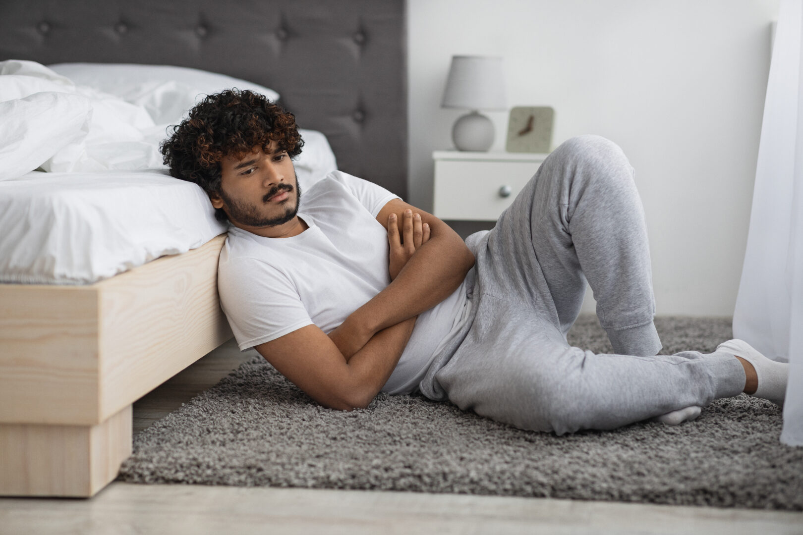 Pensive young indian man in pajamas sitting on floor by bed, thinking about something, looking for solution while COVID-19 pandemic crisis, looking at copy space, side view, panorama