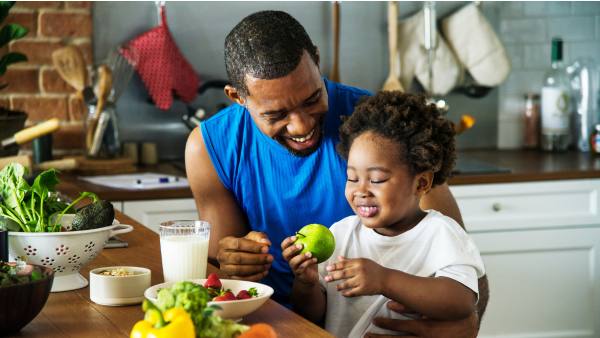 Man in blue shirt in kitchen with daughter eating fruit