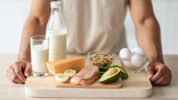 Wooden cutting board with chicken, eggs, milk, avocado, and nuts with man's arms