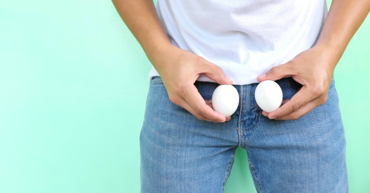 Man in front of a green wall wearing blue jeans and holding two eggs